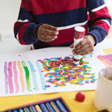 Child using a Zart Colour Apps bottle to stamp colourful dots onto white paper. Overlapping circles form a dense, textured artwork beside painted colour swatches. The child’s hands hold the bottle upright over the artwork.