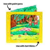 A sample artwork 3D diorama book made using Zart Card Squares White, shown from the front. A painted forest scene with two bear characters and a picnic rug sits within a yellow card frame. Two annotations read "Use with paint pens" and "Use with Zart Slicks" pointing to the artwork.