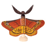 A moth-shaped sample made using a Zart Wooden Peg and Stand piece, with paper wings hand-painted in orange, yellow and brown featuring large eye-spot markings. The peg's rounded top forms the moth's head, mounted upright on its wooden stand.
