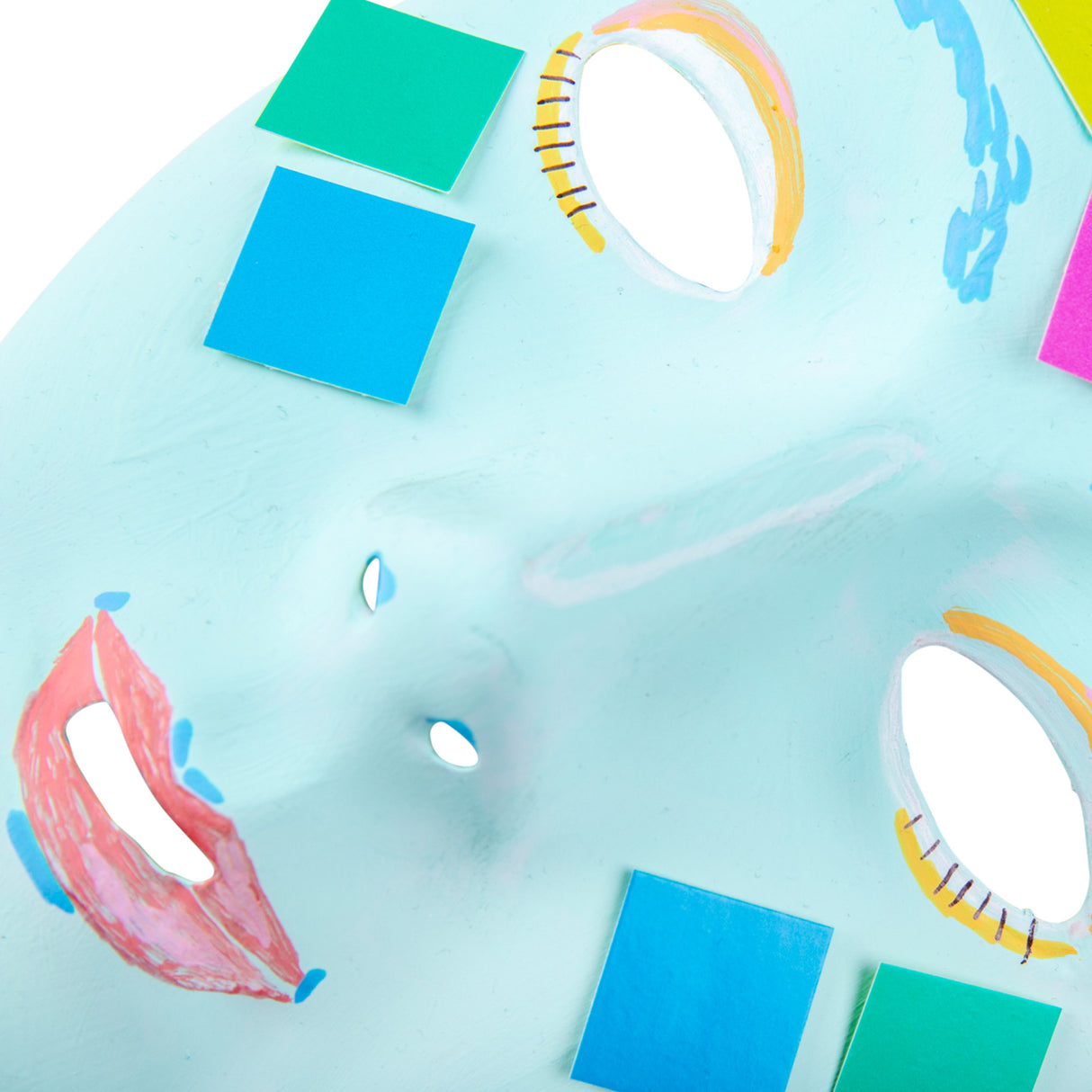 A close-up detail shot of a decorated Zart White Face Mask. The pale blue painted surface features painted eyes with yellow and orange lids and fine lash marks, red-pink lips, and applied paper squares in bold blue, teal, and green tones. 