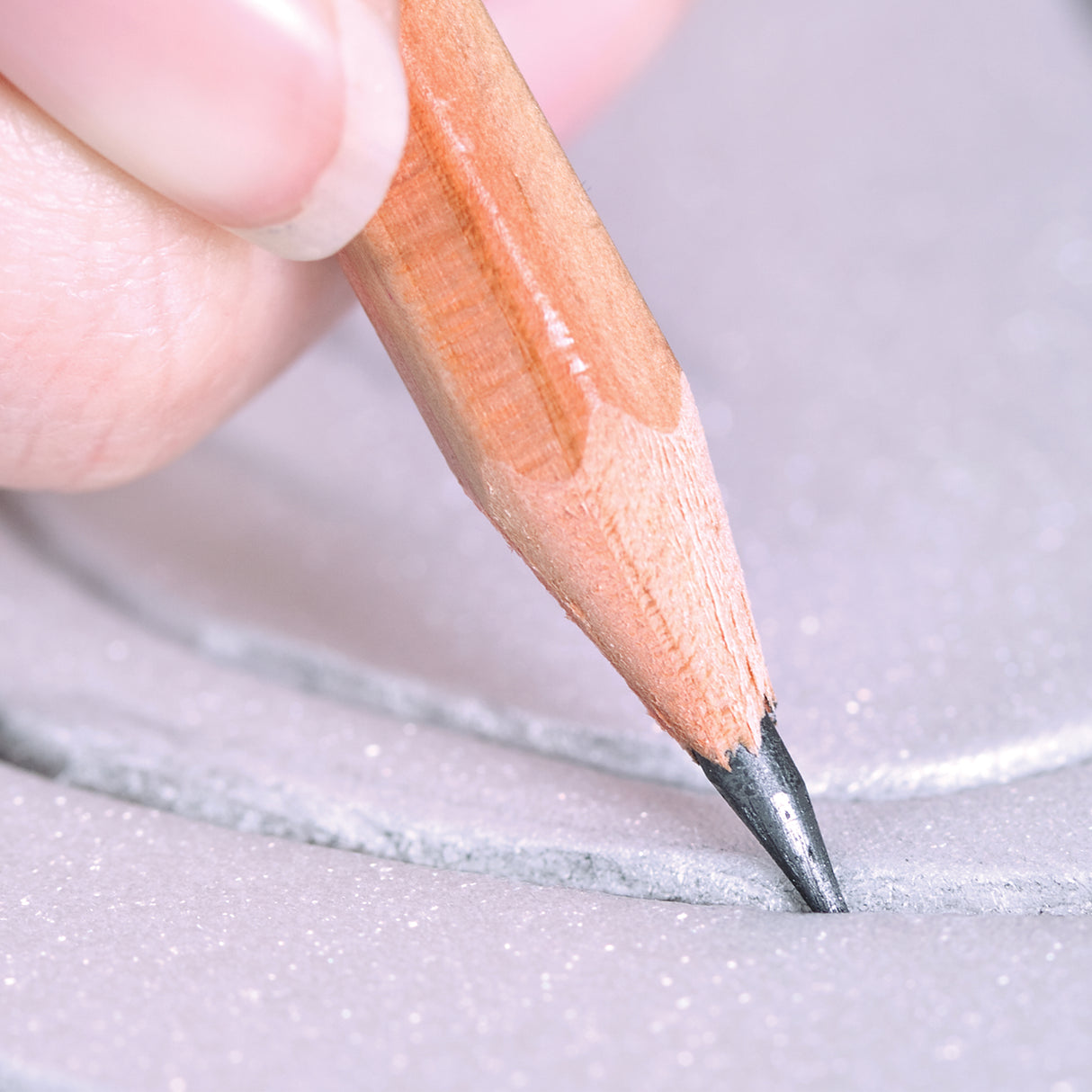 A close-up shows a hand holding a wooden pencil, its graphite tip pressed into a shallow, curved groove along the edge of a pale lilac, glitter-flecked Zart Printing Foam Round. The indented line curves beside a straight cut edge, with fine sparkling particles visible across the surface.
