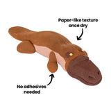 Platypus sculpture made from layered brown and beige Paper Magiclay. On-image annotations read Paper-like texture once dry and No adhesives needed, with arrows pointing to the body and limbs. Matte surface on a white background.