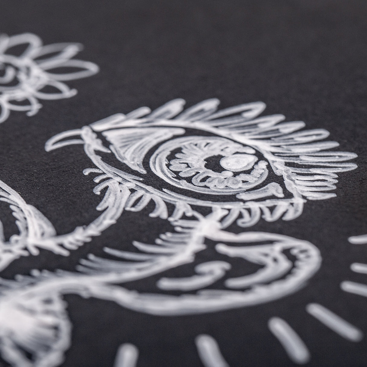 Close-up detail of intricate white line art on black paper, showing an ornate design featuring a stylised eye with decorative feathered rays and curved flourishes. The White Perma Pen by Zart creates crisp, opaque lines with excellent coverage, demonstrating fine control for detailed illustration work.