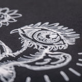 Close-up detail of intricate white line art on black paper, showing an ornate design featuring a stylised eye with decorative feathered rays and curved flourishes. The White Perma Pen by Zart creates crisp, opaque lines with excellent coverage, demonstrating fine control for detailed illustration work.