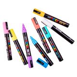 Assorted Uni POSCA pastel paint markers arranged loosely on a white background, with some caps removed to show the medium bullet tips. Colours include peach, pink, yellow, mint, blue, white and purple.