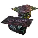 Two decorated Scratch Graduation Hats. The rear hat is covered in rainbow flowers and leaves scratched from the black surface. The front hat shows the scratched name "LINDA", "CLASS of", an owl and handwritten messages in pink, blue and green.