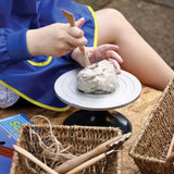 Child wearing blue art smock uses wooden modelling tool to shape grey clay on an 18cm pottery Turning Wheel, a circular metal turntable with concentric ring guides. Woven baskets containing additional tools sit nearby on sandy work surface.