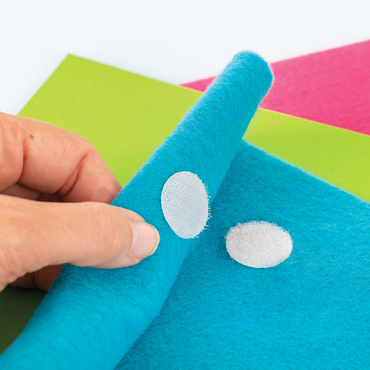 Hand placing a white loop fastener dot onto teal felt fabric, showing contrast between soft fibrous surfaces. A second dot rests nearby on the felt, with bright coloured paper sheets in the background adding smooth, bold colour contrast