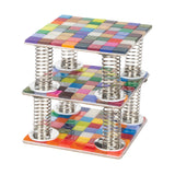 A layered sculpture of three square panels tiled with multicoloured Zart Deco tiles, stacked one above the other on coiled silver springs. Each panel carries a dense grid of glossy tiles in mixed colours — purples, greens, reds, blues and yellows — creating a bright, floating tiered effect.