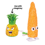 Two textured foam-covered figures, a pineapple and a tall corn-like form, are mounted on wind-up walking mechanisms. The pineapple has painted facial features and layered green leaves, while the orange corn shape has a beaded surface and wide eyes, alongside the text “Use with Magiclay.”