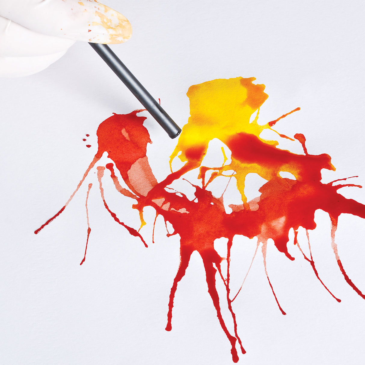 A blow-painting technique using Eco Colour Liquid Food Dyes in red and yellow, with a drinking straw shown directing splattered droplets of dye across white paper. The colours branch outward in thin, vein-like trails, creating a spontaneous, organic splatter pattern.