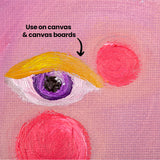 Close-up of a painted eye on pink canvas with a mustard yellow brow, purple iris and two bright pink circular forms nearby. Black annotation text reads Use on canvas & canvas boards with an arrow pointing to the painted surface, highlighting the visible woven canvas texture.