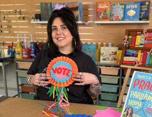 Presenter Francesca Spillane smiles while holding a bright handmade rosette featuring the word “VOTE” in bold red letters. Colourful paper ribbons spill across the table, with paint bottles, books, and classroom storage creating a vibrant studio backdrop for Zart Professional Learning Book Week 2026.