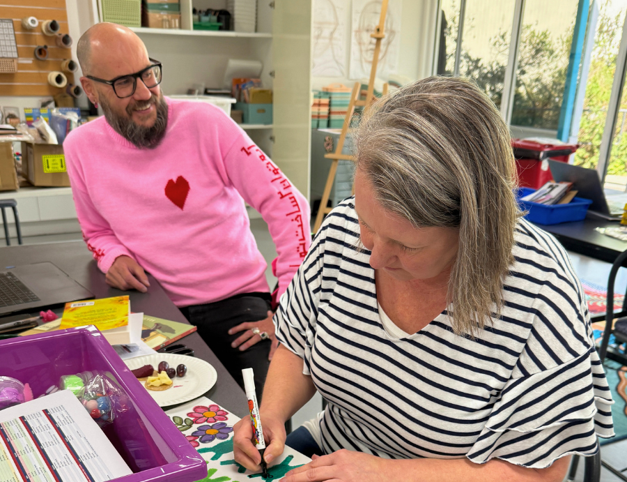 A woman draws frog designs with a marker at a table while another person in a pink jumper with a heart motif looks on, smiling. Art materials, containers, and a laptop surround them in a bright studio, documenting collaboration at Zart Professional Learning Book Week 2026.