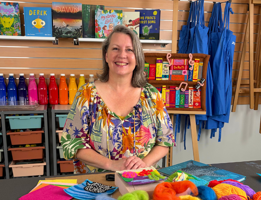 Presenter Andrea Kaltwasser smiles behind a table displaying colourful wool fibres, felt pieces, and textile tools. Bright paint bottles line the shelves behind her alongside children’s books and blue tote bags, creating a lively classroom studio scene for Zart Professional Learning Book Week 2026.