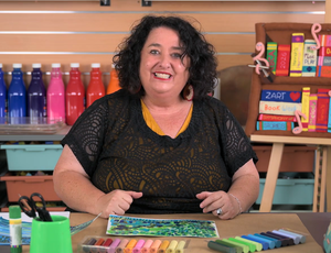 Presenter Tania Di Berardino smiles while seated at a table displaying a colourful artwork and neatly arranged pastel sticks. Bright paint bottles, classroom storage, and a book display fill the background, creating an engaging studio environment for Zart Professional Learning Book Week 2026.