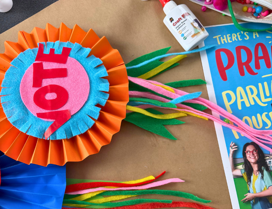 A close-up of a layered paper rosette in bright orange, blue, and pink displays the word “VOTE” in bold red letters. Colourful felt strips and craft materials surround it, alongside a glue bottle and book, highlighting hands-on making at Zart Professional Learning Book Week 2026.