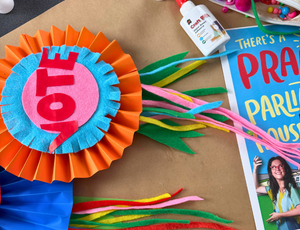 A close-up of a layered paper rosette in bright orange, blue, and pink displays the word “VOTE” in bold red letters. Colourful felt strips and craft materials surround it, alongside a glue bottle and book, highlighting hands-on making at Zart Professional Learning Book Week 2026.