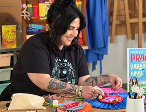 Presenter Francesca Spillane focuses on assembling a colourful paper rosette, using glue to attach small decorative elements. Bright craft materials, ribbons, and tools spread across the table, with books and classroom displays behind, documenting hands-on making at Zart Professional Learning Book Week 2026.