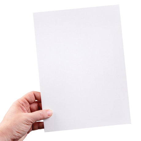 A hand holds a medium sized canvas board slightly angled against a pale background. The white primed surface shows a fine, even canvas weave. The board appears thin with straight clean edges and a firm backing.