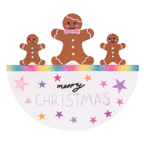 A hand-decorated Zart Rocking Christmas Card featuring a gingerbread family — one large and two small figures with white icing details — standing along a rainbow glitter edge. Below, "merry Christmas" is handwritten among pink, purple and orange foil stars scattered across the white card.