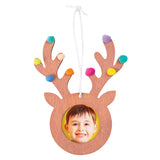 Wooden reindeer-shaped frame with white hanging cord, featuring a circular photo opening displaying a child's smiling face. The natural wood frame is decorated with colourful pom-poms in turquoise, purple, yellow, orange, pink and blue adorning each antler point, creating a festive Christmas ornament craft sample.