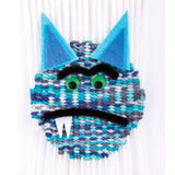 Woven yarn craft shaped like a stylised cat face in cool blue and teal tones, showing tight interlaced fibres. Features bright green googly eyes, soft felt triangular ears, and fuzzy black pipe cleaner details forming eyebrows and mouth.