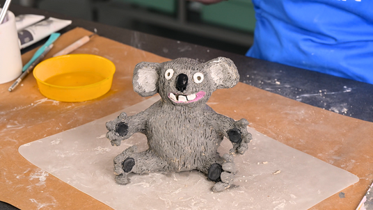 Hand-built ceramic drop bear sculpture with textured grey fur, large ears and an expressive face, displayed on a pottery work surface surrounded by clay tools and materials during the making process from the Zart Education On Demand activity 'Ceramic Drop Bears'.