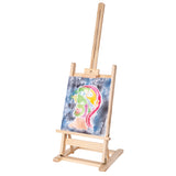 Angled view of a natural wood tabletop easel holding a colourful painted portrait artwork featuring pink, green, yellow and blue tones with soft watercolour textures. The adjustable easel frame includes brass fittings, visible woodgrain detail and a tilted support structure against a light grey background.