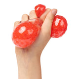 A hand squeezing three translucent red gel-filled stress balls with textured surfaces that create bumpy, organic shapes. The EmotiBalls are squishy sensory toys filled with colourful beads and liquid, demonstrating their flexible, tactile nature. The balls catch light, showing their glossy, semi-transparent material perfect for stress relief and sensory exploration.