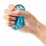 Hand squeezing a translucent blue Sensory Sprouts Jelly Cube filled with swirling turquoise and white patterns and small confetti pieces. The squishy, gel-filled sensory toy demonstrates its flexible, tactile texture as fingers compress the smooth, dome-shaped cube against a white background.
