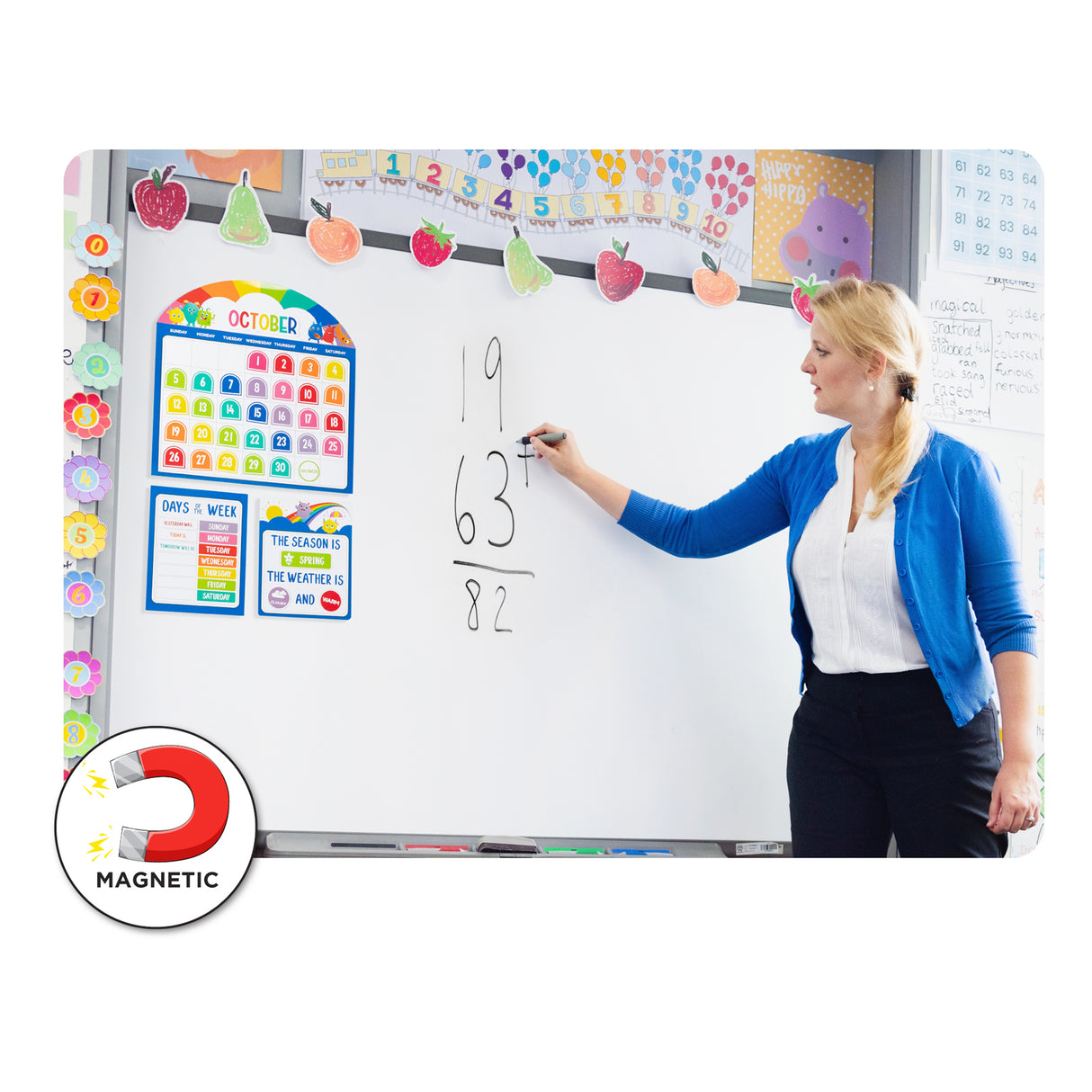 Teacher in blue cardigan writing mathematics equations on whiteboard decorated with Giggle & Grow Magnetic Calendar Bulletin Board Set featuring colourful October calendar, days of the week chart, weather tracker, and decorative fruit garland border. MAGNETIC icon displayed in bottom left corner showing magnet symbol.