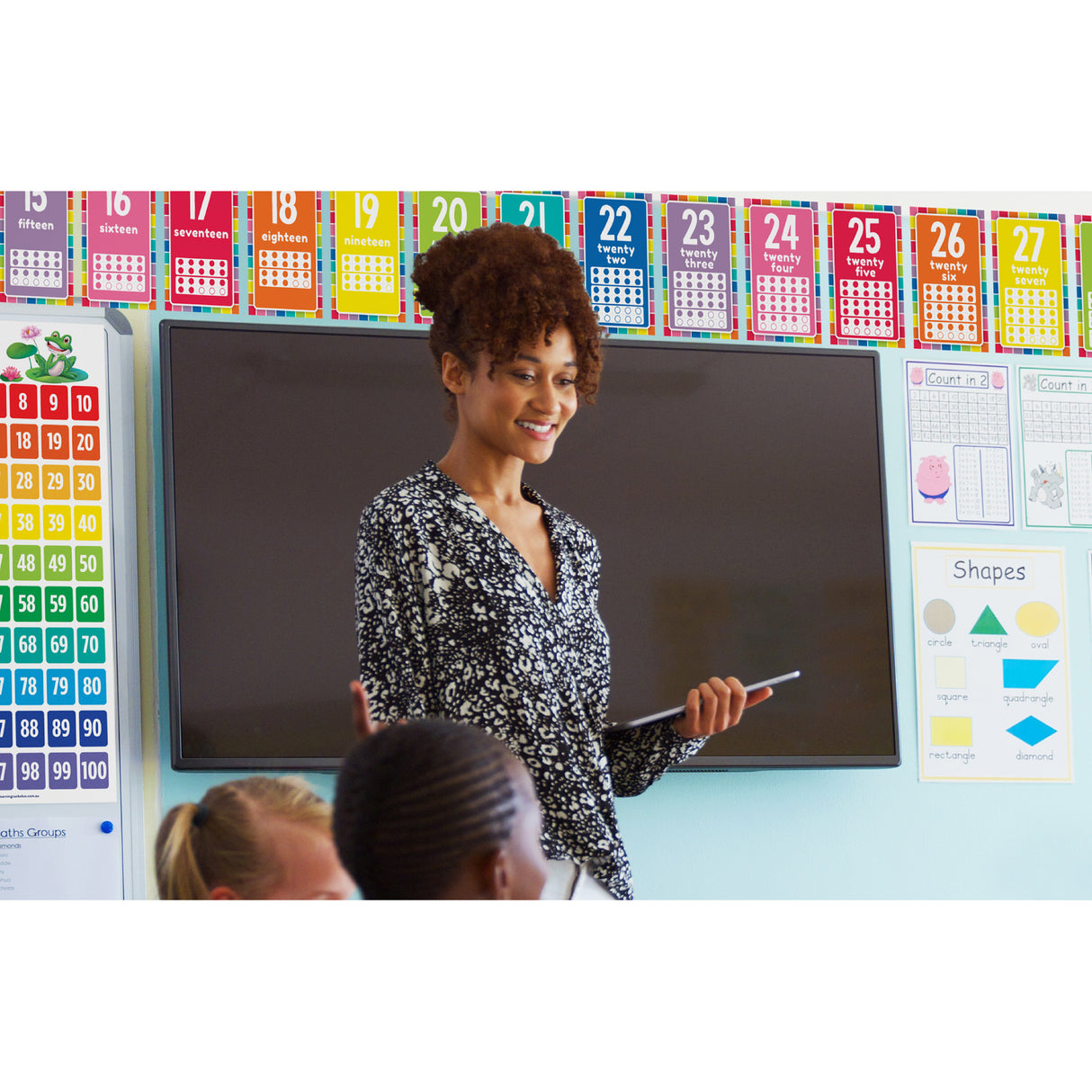 Colourful classroom display featuring number cards numbered 15 to 27 showing numerals and counting dots in vibrant colours including pink, orange, yellow, teal and blue. The bulletin board set includes additional educational posters showing counting exercises with Count in 2s worksheets and a Shapes poster displaying circle, triangle, oval, square, quadrangle, rectangle and diamond shapes with labels.