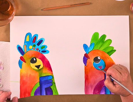 Zart Education PL Online Exploring Watercolour and Ink — A person paints two brightly coloured, stylised birds with vibrant gradients of red, orange, yellow, green, blue, and purple. The birds feature bold crests, large expressive eyes, and graphic shapes. A brush, pencil, water jar, and paper towel sit around the workspace, highlighting a playful watercolour illustration activity.