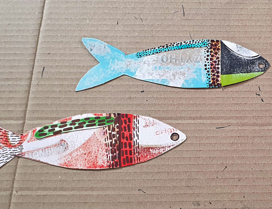 Zart Education Workshop-in-a-Box Upcycled Card and Paper close-up of two colourful fish-shaped cut-outs made from recycled printed card. The top fish features turquoise, black, and lime-green sections with dotted patterns, while the bottom fish combines red, brown, and green textured areas. Both pieces rest on a corrugated cardboard background, showcasing creative reuse of materials.