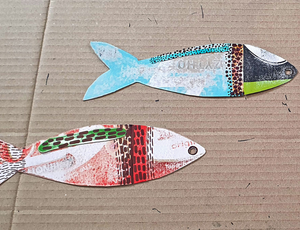 Zart Education Workshop-in-a-Box Upcycled Card and Paper close-up of two colourful fish-shaped cut-outs made from recycled printed card. The top fish features turquoise, black, and lime-green sections with dotted patterns, while the bottom fish combines red, brown, and green textured areas. Both pieces rest on a corrugated cardboard background, showcasing creative reuse of materials.