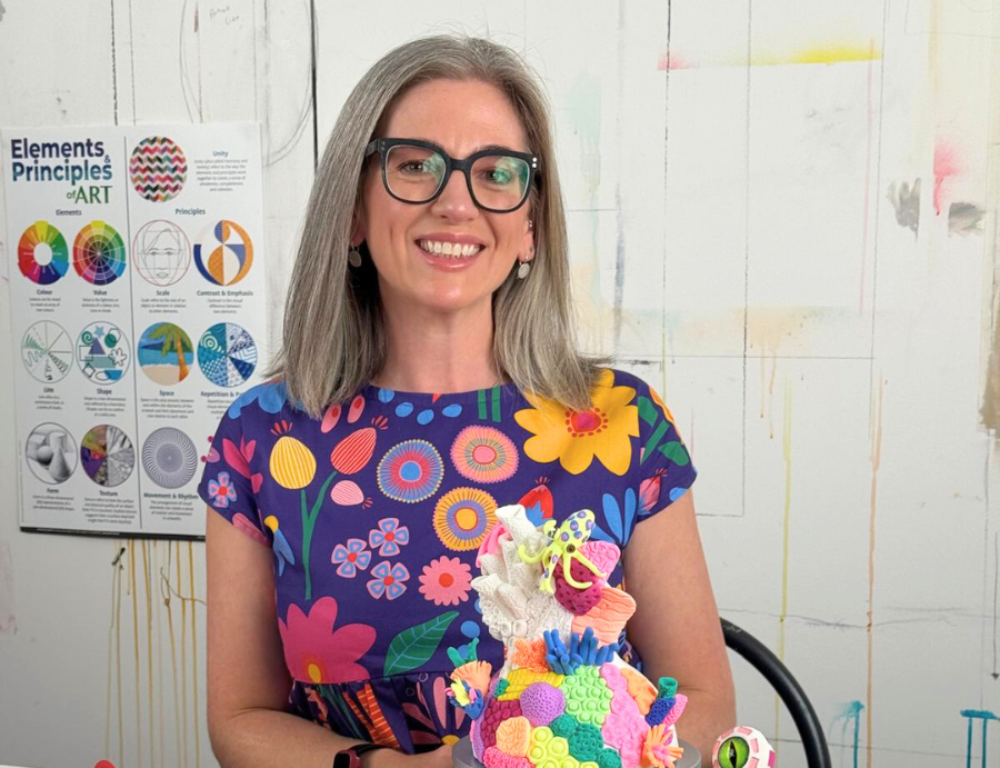 Dee Zabel, presenter of Zart Education PL “The Essentials of Modelling”, smiling and seated at a studio table holding a colourful mixed-media sculpture on a black display stand. She wears black glasses and a vibrant floral dress. The table is covered with modelling materials including tubs of clay and foam beads, sculpting tools, sketches, and small sculptural examples such as a burger model and mosaic-patterned creatures. An “Elements & Principles of ART” poster is visible on the wall behind her in an art 