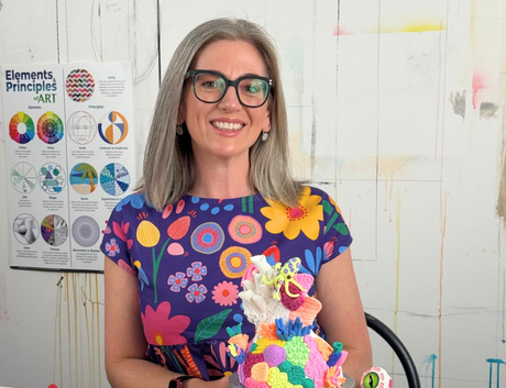 Dee Zabel, presenter of Zart Education PL “The Essentials of Modelling”, smiling and seated at a studio table holding a colourful mixed-media sculpture on a black display stand. She wears black glasses and a vibrant floral dress. The table is covered with modelling materials including tubs of clay and foam beads, sculpting tools, sketches, and small sculptural examples such as a burger model and mosaic-patterned creatures. An “Elements & Principles of ART” poster is visible on the wall behind her in an art 