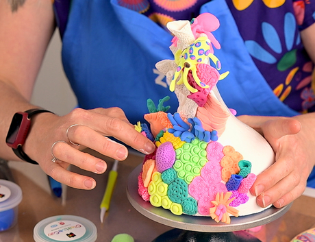 Close-up photo of hands holding a colourful sculptural model on a turntable. The sculpture is covered in bright textured pieces in pink, yellow, green, blue and orange. Art materials and small tubs sit nearby on the table. Credit: Zart Education PL The Essentials of Modelling.