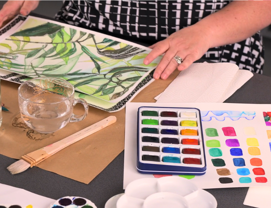 Zart Education PL Online Exploring Watercolour and Ink. Close-up of a person working on a watercolour painting of green leaves. The workspace includes a paint palette, a glass of water, a large brush, and a colour test sheet showing a range of mixed watercolours. The artist wears a black-and-white patterned top, with paper towels and painting tools neatly arranged on the table, capturing a calm creative process.