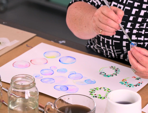 Zart Education PL Online Exploring Watercolour and Ink — A person paints delicate watercolour studies, including translucent bubbles in soft blues, pinks, and yellows, and small botanical wreaths made from fine brushstrokes. Jars of water, a mug, and brushes surround the workspace, highlighting a focused and experimental painting session.