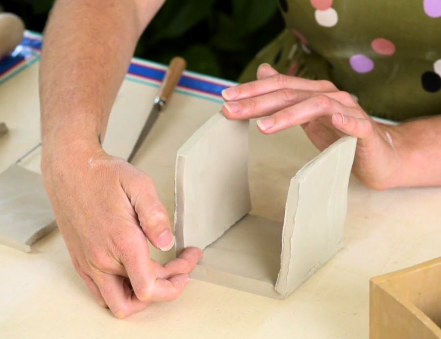 Zart Education Professional Learning The Essentials of Clay image showing hands positioning upright pale grey clay slabs onto a square clay base. The smooth slab surfaces create clean geometric forms with visible cut edges, while a clay knife, spare slab pieces and timber mould are arranged nearby.