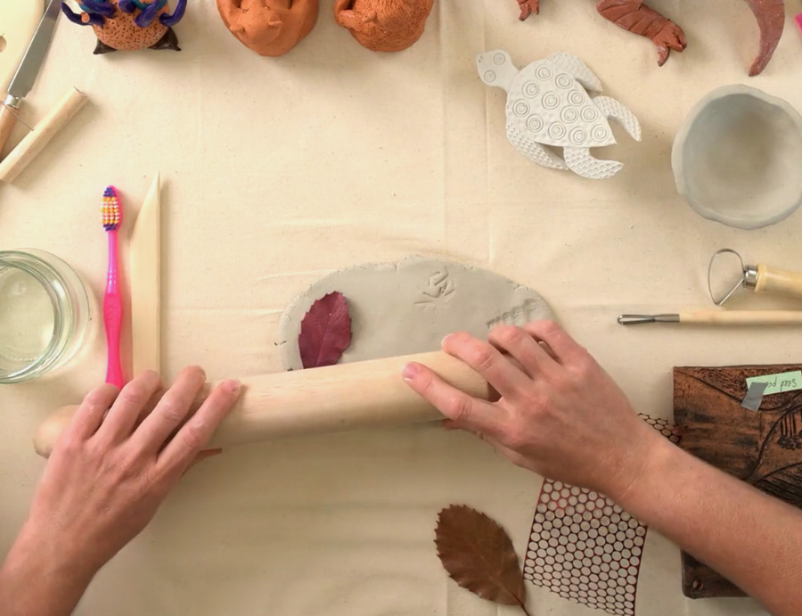 Zart Education Professional Learning The Essentials of Clay image showing hands rolling a wooden rolling pin across a pale grey clay slab. Leaves, textured materials, carved blocks, clay tools, a water jar and ceramic samples surround the workspace, highlighting varied surface patterns, textures and natural forms.