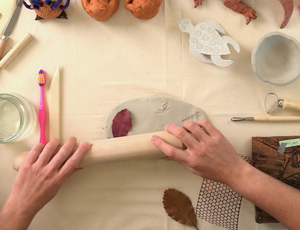 Zart Education Professional Learning The Essentials of Clay image showing hands rolling a wooden rolling pin across a pale grey clay slab. Leaves, textured materials, carved blocks, clay tools, a water jar and ceramic samples surround the workspace, highlighting varied surface patterns, textures and natural forms.