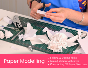 Workshop tile showing hands cutting white paper pieces over a green cutting mat. Paper shapes and partially constructed paper forms are scattered across the table with a stapler and tools nearby. Pink banner text reads “Paper Modelling” with dot points: “Folding & Cutting Skills”, “Joining Without Adhesives”, “Constructing 3D Paper Structures”. Credit: Zart Education PL The Essentials of Modelling.