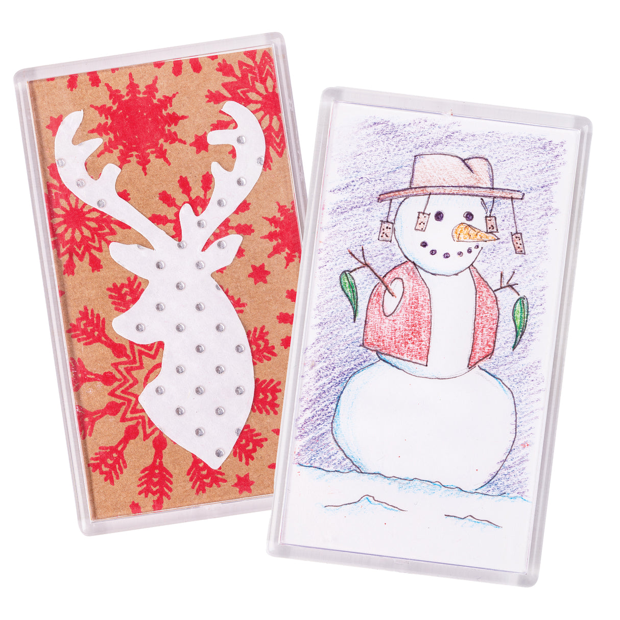 Two Zart Magnet Clear Frames stand side by side, each holding a decorated card behind clear plastic. The left frame shows a white reindeer head with silver dot detailing on a red snowflake-patterned kraft background. The right frame holds a coloured pencil snowman wearing a hat, scarf, and mitten-shaped gift tags.