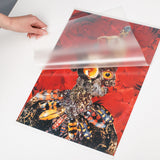 A hand lifts a translucent Zart Laminating Pouch above a vivid insect artwork with red floral tones and patterned beetles. The frosted pouch surface partially softens the image beneath while showing the flexible protective plastic texture and smooth semi-transparent finish.