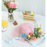 Pink woven straw Easter bonnet decorated with colourful eggs, flowers, ribbons and ornamental chicks, displayed on white table alongside craft decorations including felt chicken ornaments, glass vase with fresh flowers, and wooden crate filled with Easter craft supplies.