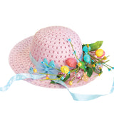 Pink woven straw bonnet decorated with a light blue satin ribbon, small artificial flowers in blue and yellow, miniature Easter eggs in pastel pink, yellow and turquoise, green leaves, and decorative spring embellishments around the hatband.