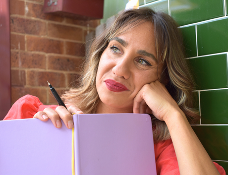 Portrait of Kirli Saunders (OAM), a proud Gunai Woman and award-winning multidisciplinary artist, seated indoors and holding a pen and notebook while gazing thoughtfully to the side.